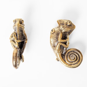 May include: Two gold-colored chameleon figurines. The chameleon on the left is curled up with its tail wrapped around its body. The chameleon on the right is curled up with its tail curled into a spiral.