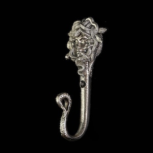 May include: A silver-coloured wall hook depicting Medusa's face with snakes. The hook is shaped like a snake, with a curved end. The metal hook has a detailed design and is suitable for hanging items.