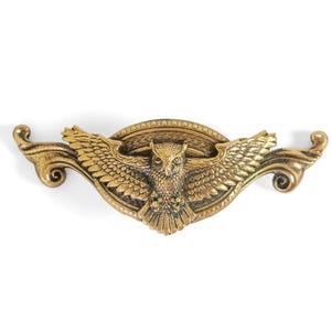 May include: A gold-colored metal owl-themed drawer pull. The owl is centered with outstretched wings, set within an oval frame. The pull has decorative scrollwork on each end, creating a symmetrical design. The hardware is likely for furniture.