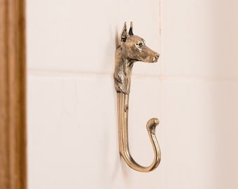 Könnte beinhalten: Ein goldfarbener Haken aus Metall mit einem Hundekopfdesign. Der Haken ist an einer weißen gekachelten Wand montiert.