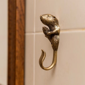 Könnte beinhalten: Ein goldfarbener Haken aus Metall in Form eines Bibers, der einen Zweig im Maul hält. Der Haken ist an einer weißen gekachelten Wand befestigt.