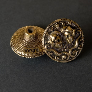 Pode incluir: Botão de metal dourado com um design em relevo de dois rostos e a palavra "TWINS" inscrita na parte inferior. O botão está preso a uma base redonda de metal dourado menor.