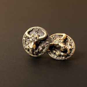 Op de afbeelding: Twee bronskleurige manchetknopen met een gedetailleerd stierontwerp. Elke manchetknop heeft een cirkelvorm met een verhoogde stierenkop in het midden, omringd door ingewikkelde details. De achtergrond is donkerbruin.