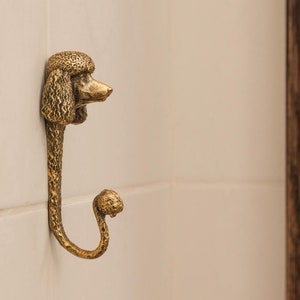 May include: A gold-toned metal hook shaped like a poodle's head. The hook is mounted on a white tiled wall.