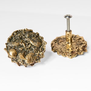 Op de afbeelding: Twee gouden kastknoppen met een getextureerd, organisch ontwerp. Eén knop heeft een zilveren schroef.