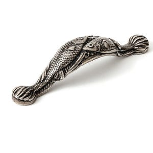 Op de afbeelding: Een zilverkleurige metalen meubelgreep met een gedetailleerd, sculpturaal ontwerp van twee vissen die in tegengestelde richtingen zwemmen. De handgreep heeft een getextureerde, antieke afwerking met zichtbare schubben, vinnen en ogen.