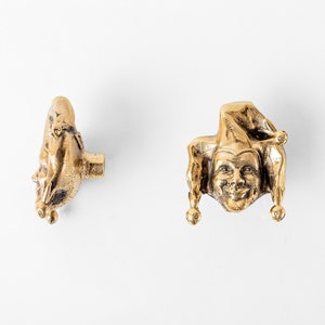 May include: Two gold-toned metal jester figurines. The jester on the left is standing on its toes with its arms raised above its head. The jester on the right is wearing a pointed hat and has a mischievous grin.