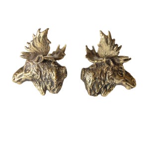 May include: A pair of brass-toned moose head earrings. Each earring depicts a detailed moose head with antlers, featuring a textured finish. The earrings are set against a white backdrop, emphasizing their intricate design and rustic appeal.