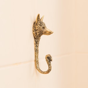 Puede incluir: Un gancho de pared de cabeza de zorro de tono dorado con un diseño detallado. El gancho tiene la forma de la cabeza y el cuerpo de un zorro, con una cola curva que forma el gancho.