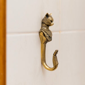 Puede incluir: Un gancho de metal dorado con forma de gato con un diseño detallado. El gancho está montado en una pared de azulejos blancos.