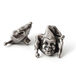 May include: Two silver-toned metal beads shaped like jester heads. Each bead features a detailed face with a smiling expression, wearing a multi-pointed hat with small spherical accents at the tips. The metal has a dark, oxidized finish.