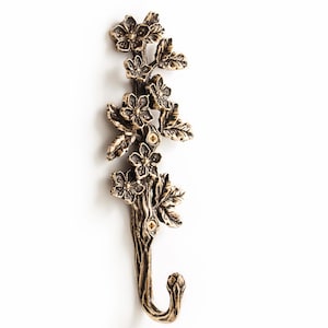 Puede incluir: Un gancho de pared vertical hecho de metal en tono bronce antiguo, con un diseño en relieve detallado de una rama floreciente con múltiples flores de cinco pétalos y hojas texturizadas. El gancho se curva hacia arriba en la base y tiene dos orificios.