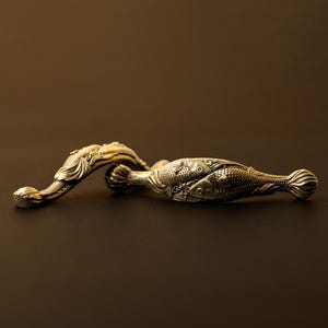 Puede incluir: Un abridor de botellas plateado y ornamentado con un diseño de pez. El abridor presenta tallas detalladas de escamas y aletas de pez, con un mango curvo. El diseño es simétrico, con los peces mirando en direcciones opuestas. El abridor es probablemente de metal.