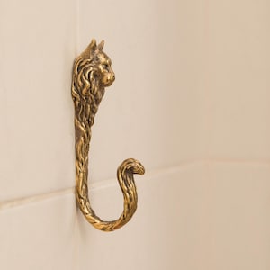 May include: A gold-toned metal hook shaped like a cat with a curled tail. The hook is mounted on a white tiled wall.