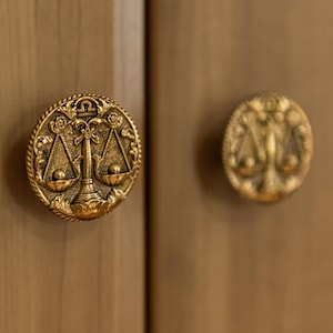 Könnte beinhalten: Zwei goldfarbene Schrankknöpfe mit einem Waage-Design. Die runden Knöpfe weisen aufwendige Details auf, darunter ein Waage-Symbol und florale Akzente. Sie sind an einer hellbraunen Holzschranktür angebracht.