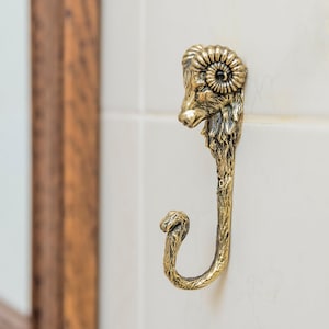 Puede incluir: Un gancho de metal dorado con forma de cabeza de carnero con cuernos rizados. El gancho está montado en una pared de azulejos blancos.