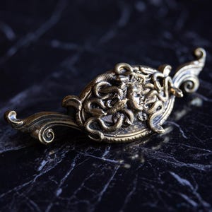 May include: A detailed, antique-style bronze Medusa brooch. The brooch features a central oval design with the face of Medusa surrounded by snakes. The bronze has a slightly aged appearance, with intricate details and a decorative, curved handle.
