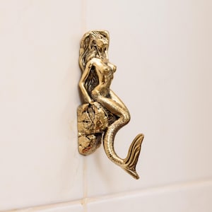 May include: A gold-toned mermaid figurine with a long tail, sitting on a rock, is used as a hook. The mermaid is facing to the right and has long flowing hair.