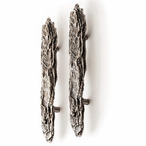 May include: Two vertical cabinet pulls with a dark silver, metallic finish. Each handle features a highly textured, rugged surface that mimics the appearance of natural tree bark, with deep crevices and raised ridges throughout the length.