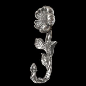 May include: A silver-toned metal wall hook shaped like a flower on a stem. The design features a single bloom at the top, textured leaves along the central stalk, and a curved bottom section that forms the hook for hanging items on a wall surface.