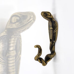Puede incluir: Un gancho de metal dorado con forma de serpiente con la cabeza levantada y la cola enroscada en un gancho. La serpiente está detallada con escamas y tiene un aspecto realista.