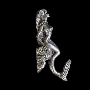 Könnte beinhalten: Eine silberfarbene Meerjungfrau-Skulptur mit langem, fließendem Haar sitzt auf einer strukturierten Felsformation. Die Meerjungfrau blickt nach oben, ihr Schwanz krümmt sich anmutig. Der Hintergrund ist tiefschwarz.