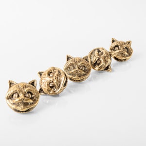 May include: Four gold-coloured metal buttons featuring a grinning cat face design. The buttons are arranged in a row, with the cat faces facing the viewer.