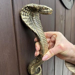Könnte beinhalten: Ein bronzefarbener Türgriff in Form einer Kobra. Der Griff ist mit Schuppen detailliert und hat den Kopf der Schlange oben. Der Griff ist an einer braunen Holztür befestigt.
