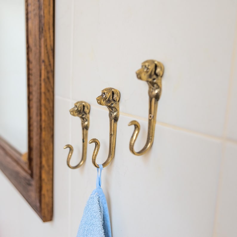 May include: Three gold-toned dog head hooks mounted on a white tiled wall. A light blue towel is hanging from one of the hooks.