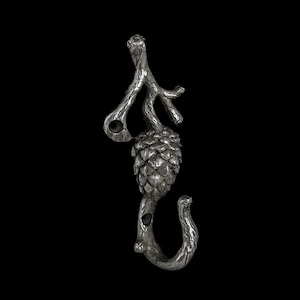 May include: A silver-toned metal hook with a pinecone design. The hook features a branch-like top with two holes and a curved bottom. The pinecone is textured, and the background is black.
