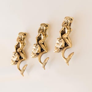 May include: Three gold mermaid hooks with a shell design. The mermaids are sitting on a shell and have their tails curled around the hook.