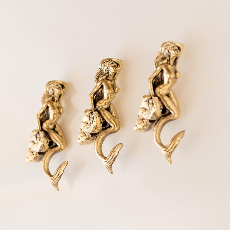 May include: Three gold mermaid hooks with a shell design. The mermaids are sitting on a shell and have their tails curled around the hook.