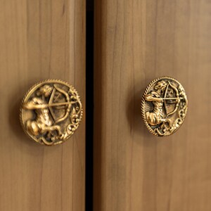 以下が含まれることがあります： 射手座のデザインが施された、2つの金色のキャビネットノブ。各ノブは、弓と矢を持ったケンタウロスを特徴とし、装飾的なボーダーで囲まれています。ノブは、明るい茶色の木製キャビネットドアに取り付けられています。