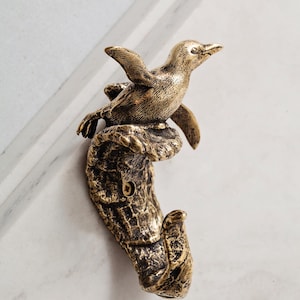 Puede incluir: Un gancho de pared hecho de metal con tono de latón antiguo, que presenta una figura de pingüino esculpida sobre una base texturizada en forma de ola. El diseño incluye un orificio para tornillo y un gancho curvo para colgar objetos.