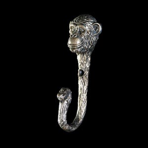 May include: A silver-coloured wall hook in the shape of a monkey. The hook has a detailed monkey head, a textured stem, and a curved hook at the bottom. The metal hook is set against a black background.