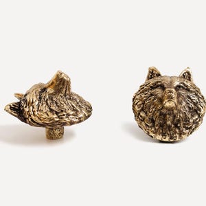 May include: Two gold-toned metal wolf head drawer pulls. The wolf head on the left is facing upwards, while the wolf head on the right is facing forward.