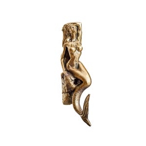 May include: A gold-coloured mermaid figurine sitting on a rock with her tail curled around her. The mermaid is depicted with long flowing hair and a serene expression.