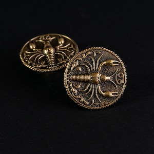 Peut inclure: Deux boutons dorés antiques avec un motif détaillé du signe astrologique du Cancer. Chaque bouton présente un crabe au centre, entouré de volutes ornées et d'une bordure en forme de corde. Les boutons sont posés sur un fond sombre.