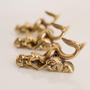 May include: Four gold-toned mermaid figurines with tails curled up, each resting on a rock-like base. The mermaids are arranged in a row, with the tails facing the viewer.