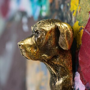 May include: A gold-colored metal sculpture of a dog's head, with the dog's head turned to the side. The sculpture is mounted on a colorful background.