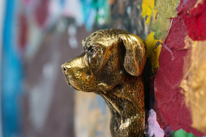 May include: A gold-colored metal sculpture of a dog's head, with the dog's head turned to the side. The sculpture is mounted on a colorful background.