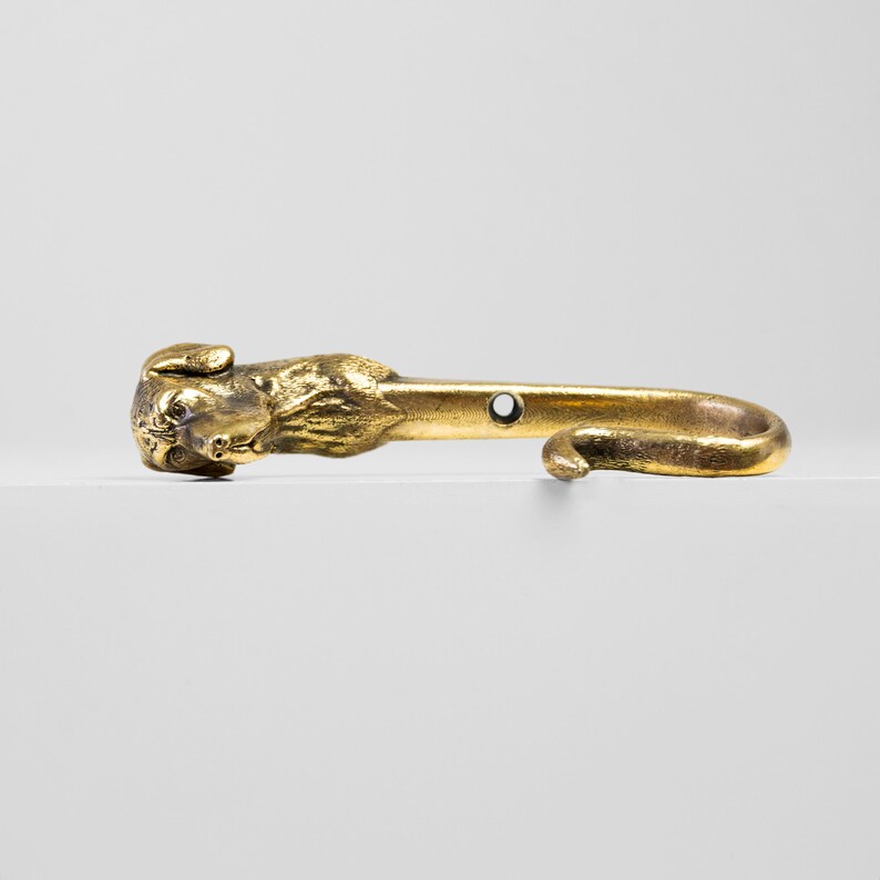 May include: A gold-toned metal hook with a dog's head at the top. The hook is curved and has a hole for mounting.