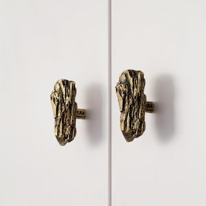 May include: Two gold-toned cabinet knobs with a wood-like texture. The knobs have a rustic, natural look.