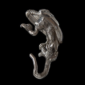 Pode incluir: Um gancho de metal prateado em forma de uma águia estilizada pousada num galho. A águia tem penas detalhadas e um bico curvo. O gancho foi projetado para pendurar objetos e está sobre um fundo preto.