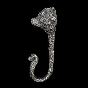 May include: A silver-coloured, J-shaped hook with a bear head at the top and a textured, tree-like stem. The hook curves at the bottom, featuring a smaller, stylised animal head. The background is solid black.