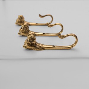 Op de afbeelding: Drie gouden haken met dierlijke kopontwerpen. De haken zijn gemonteerd op een wit oppervlak.