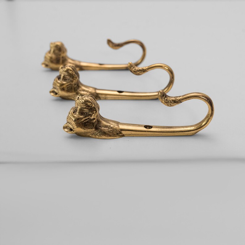 Op de afbeelding: Drie gouden haken met dierlijke kopontwerpen. De haken zijn gemonteerd op een wit oppervlak.