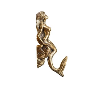 May include: A gold-toned mermaid figurine with a long tail, sitting on a rock, with her tail curled up to form a hook.