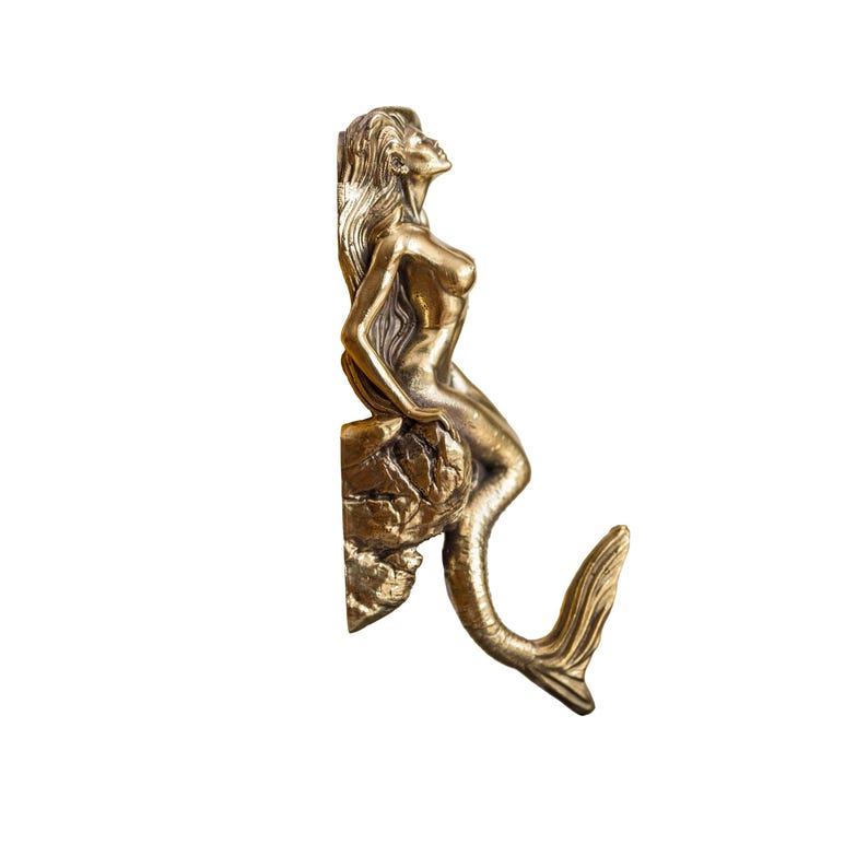 May include: A gold-toned mermaid figurine with a long tail, sitting on a rock, with her tail curled up to form a hook.