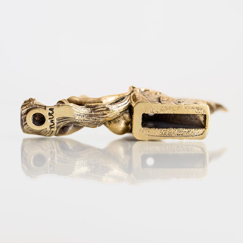 May include: A gold-coloured metal sculpture of a woman with a long flowing dress. The sculpture is mounted on a rectangular base with a slot on the bottom. The base is marked with the word "TUVUCA".